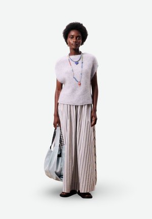 Femme portant un haut en tricot sans manches lavande, un pantalon à jambe large rayé, des colliers colorés en perles, des sandales noires, et portant un grand sac gris.