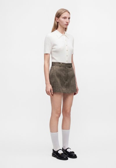 Femme debout portant une chemise blanche à manches courtes boutonnée, une mini-jupe marron à fines rayures avec boucle, des chaussettes blanches et des mocassins noirs.
