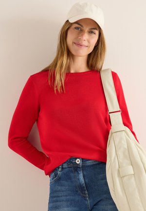 Röd långärmad tröja med rund halsringning, i kombination med blå jeans och en krämfärgad axelväska. Kappen är off-white och har en enkel design.
