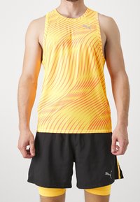Sportovní tílko Puma v jasně žluté barvě s červeným vlnkovým vzorem, doplněné černými šortkami a žlutými kompresními šortkami pod nimi.