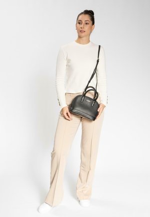 Bolso de mano de cuero negro con acabado texturizado, diseño redondeado, doble asa y correa ajustable, sostenido por una persona que lleva una blusa blanca y pantalones beige.