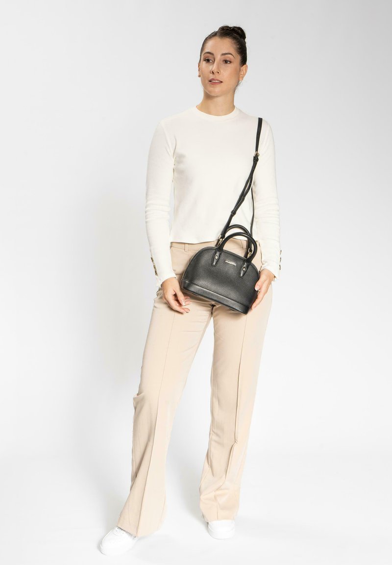 Borsa a tracolla nera in pelle con finitura texturizzata, design arrotondato, doppi manici e tracolla regolabile, tenuta da una persona che indossa una maglietta bianca e pantaloni beige.
