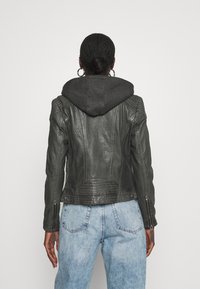 Vrouw met gevlochten haar in een lage bun, gekleed in een donkergrijze leren jack met capuchon en lichtblauwe jeans, met de rug naar de camera gericht tegen een effen achtergrond.