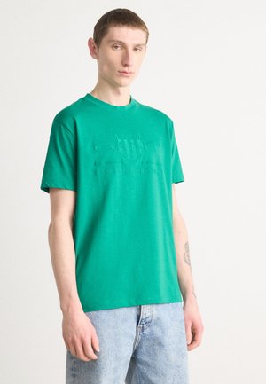Grønn bomull T-skjorte med korte ermer, med opphøyd logo på brystet. Sammen med lyseblå denimjeans. Enkel design.