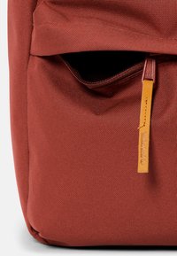 Röd textilryggsäck med en framficka, strukturerad tyg och en brun läderetikett fäst vid dragkedjan.