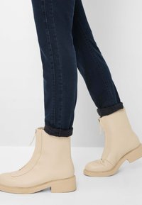 Beige læder ankelsko med lynlås, firkantet tådesign og en chunky hæl. Båret med mørke jeans rullet ved åbningerne.