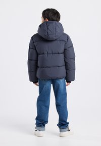 Tummanharmaa puhvijakku hupulla, jossa on tikattu muotoilu. Yhdistetty sinisiin farkkuihin ja valkoisiin lenkkareihin, nähtynä takaa.