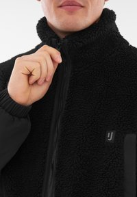 Giacca in fleece nera con alto colletto, chiusura con zip e superficie testurizzata. Presenta una piccola toppa con logo sul davanti e polsini elasticizzati.