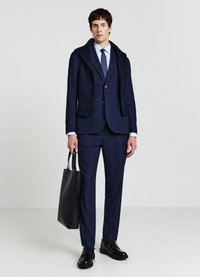 Completo blu navy con felpa con cappuccio in maglia, tessuto strutturato, pantaloni a fantasia e scarpe in pelle nera. Tiene in mano una borsa tote nera.