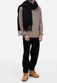 Maglione beige a coste, sciarpa nera con frange, pantaloni neri e stivali color sabbia. I materiali sembrano lavorati a maglia e morbidi, con una vestibilità casual.