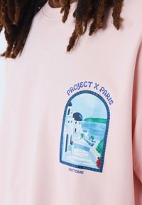 Sudadera de color rosa claro con un gráfico colorido de Santorini que presenta una escena arquitectónica en blanco y azul. El texto dice "PROJECT X PARIS" y "SANTORINI."