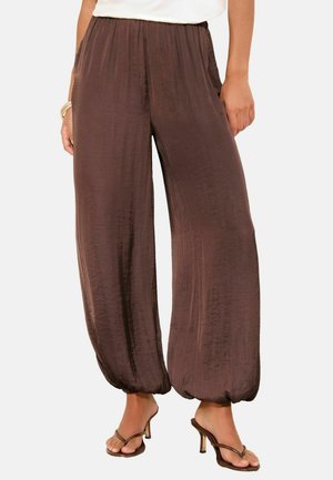 Pantalon marron ample en soie avec chevilles élastiques froncées, porté avec des sandales marron à talons et un haut blanc, les mains reposant négligemment sur les côtés.