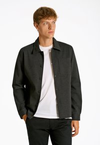 Mörkgrå knapp-up jacka i ett mjukt tyg, med krage, framficka och en avslappnad passform över en vit t-shirt.