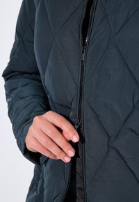 Giacca imbottita trapuntata di colore blu scuro con zip frontale. Il tessuto ha una texture liscia e presenta motivi di cucitura a forma di diamante.