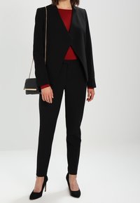 Schwarzer, taillierter Blazer und schmale Hose über einem roten Top, kombiniert mit schwarzen High Heels und einer kleinen Umhängetasche mit goldener Kette.