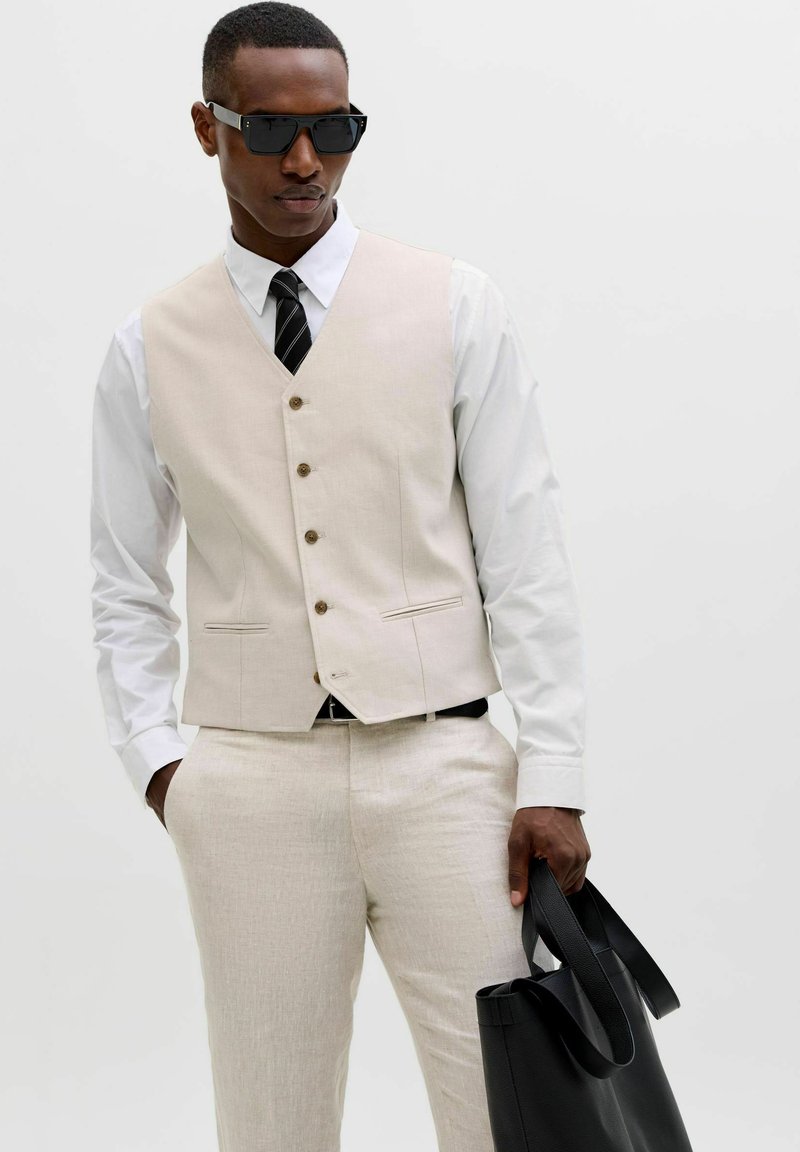 Homme portant un gilet beige et un pantalon avec une chemise blanche et une cravate noire, tenant un sac en cuir noir et portant des lunettes de soleil noires.