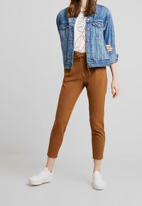 Chaqueta de mezclilla de color azul claro con dos bolsillos frontales, combinada con pantalones ajustados marrones y zapatillas blancas. Textura suave, diseño casual.