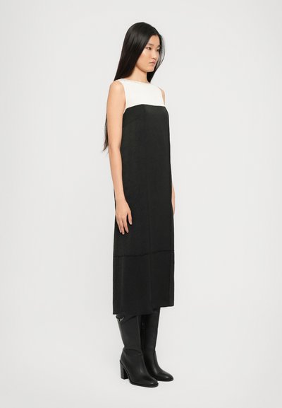 Naine seisab ilma varrukateta mustas kleidis, millel on valge ülemine paneel, seljas mustad kõrged kontsaga põlvekotid, taust on ühtlane.