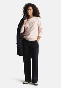 Felpa rosa chiaro con logo "GANT", abbinata a pantaloni larghi in denim scuro e sneaker beige. Un cappotto blu marino è appoggiato sulla spalla.