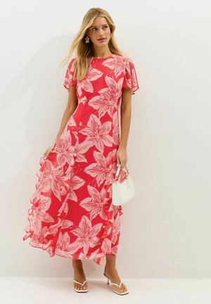 Femme portant une robe rouge fluide à grand imprimé floral rose clair, tenant un petit sac à main blanc et portant des sandales blanches à talons.