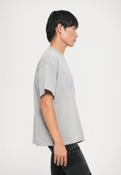 Lys grå, oversized T-skjorte med korte ermer, rund hals og lilla grafikk på forsiden; har en subtil design ved kanten.