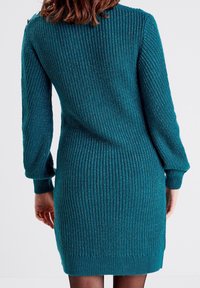 Robe en maille côtelée turquoise avec manches longues, col rond, tissu texturé et ourlet légèrement évasé. Présente des accents de fil métallique.