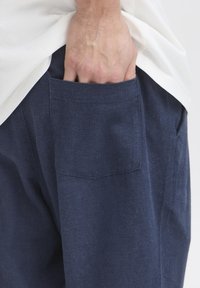 Tmavě modré lněné kalhoty s texturovaným povrchem, vybavené zadní kapsou a rukou vloženou dovnitř, doplněné bílou košilí.