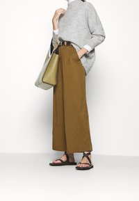 Šedý pletený svetr, hnědé kalhoty s širokými nohavicemi, černé sandály se studny a velká oliově zelená taška s černými akcenty.