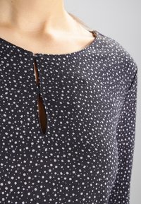 Svart topp med små vita prickar; har en rund halsringning och en nyckelhålsutskärning fram. Mjuk, texturerad tyg.