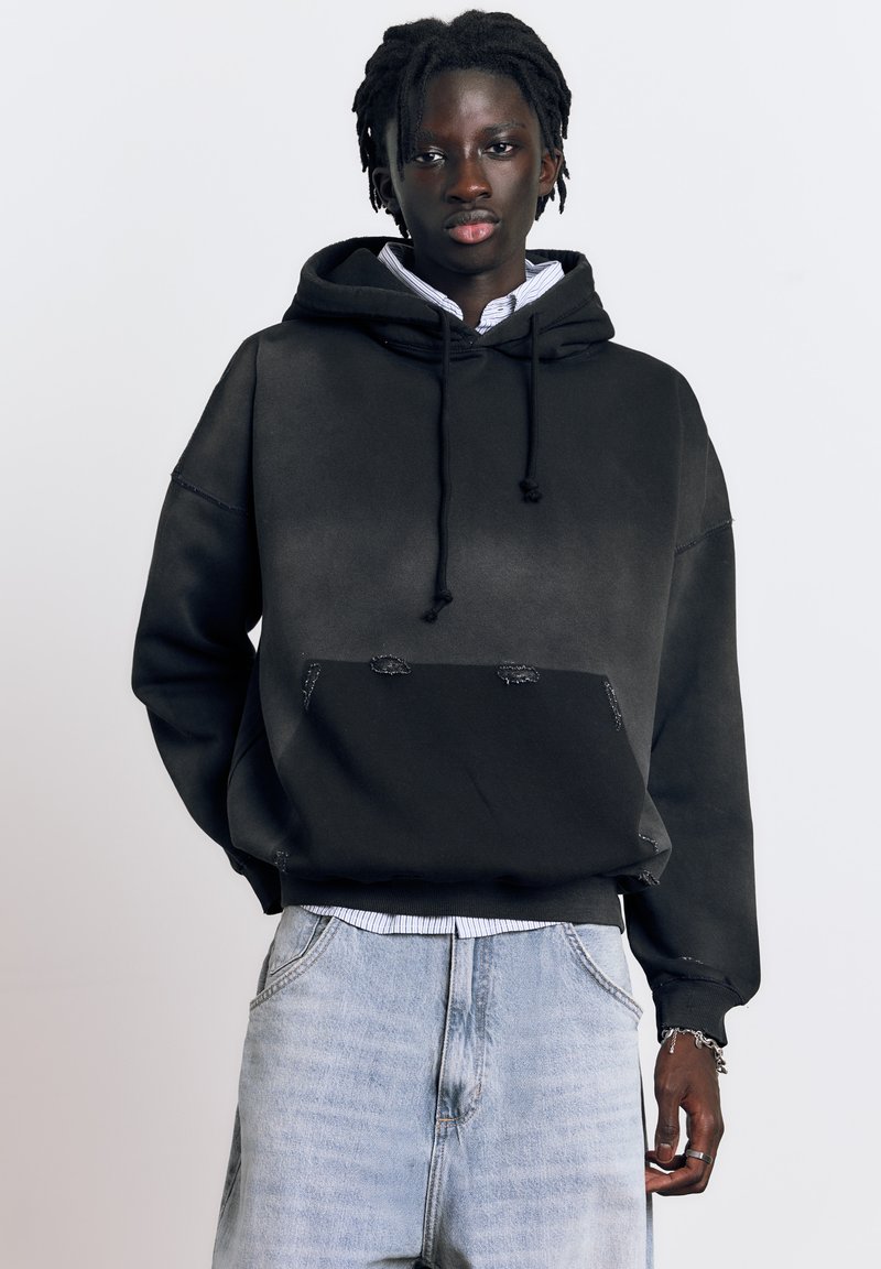 Fiatal, sötét bőrű személy dreadlock hajjal, fekete kapucnis pulóvert visel, alatta csíkos ing, világoskék farmer és ezüst ékszerek a jobb csuklóján és kezén.