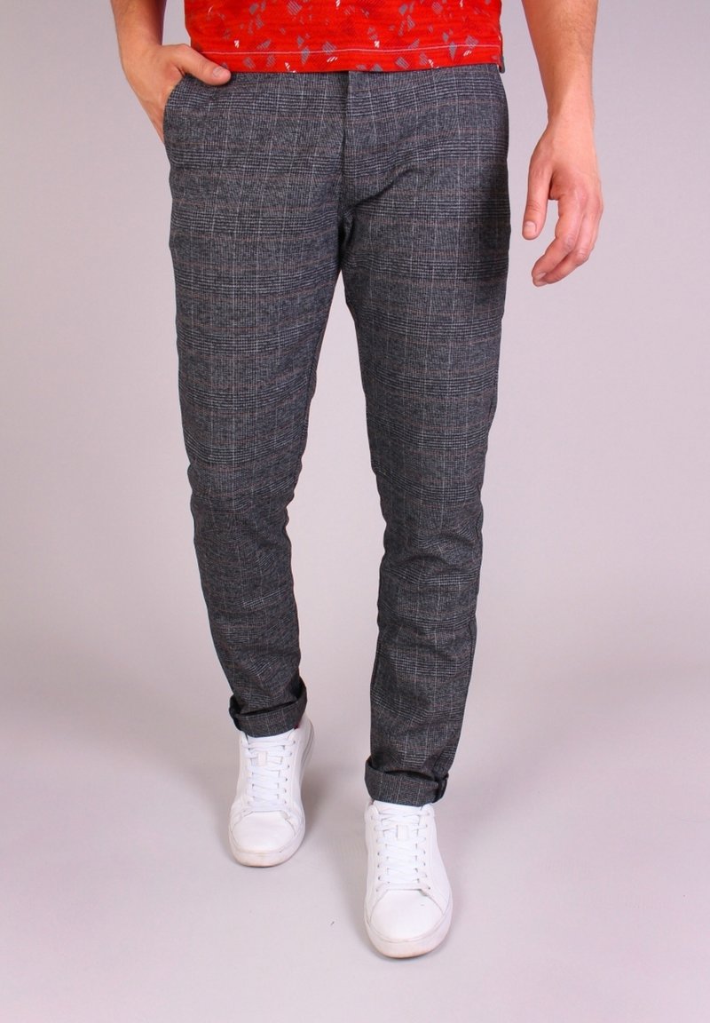 Homme portant un pantalon gris à carreaux, des baskets blanches et une chemise rouge, avec une main dans la poche, debout devant un fond uni.