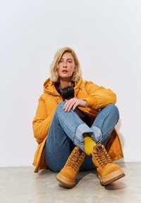 Gele gewatteerde jas, denim jeans, bruine sweater, gele sokken en bruine veterboots tot de enkel. De vloer is van beton en de achtergrond is wit.