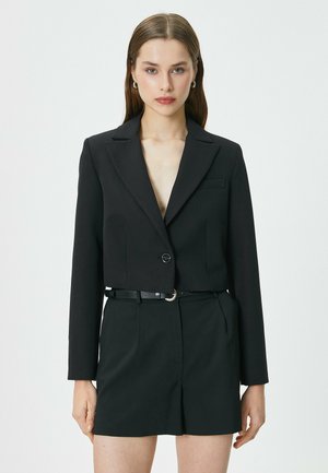 Zwarte cropped blazer met een enkele knoopsluiting, aangeknipte revers en een gladde stof; gecombineerd met hoge zwarte shorts.