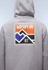 Grijze hoodie met een grote, kleurige grafiek op de achterkant met de tekst "NAPAPIJRI 87," geometrische vormen in oranje, blauw en zwart.