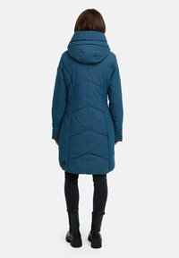 Lang, polstret kåpe i dyp blå med stor hette, sildebensmønster og logodetalj ved kanten, kombinert med svarte leggings og støvler.