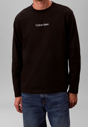 Mann trägt schwarzes Langarmshirt mit weißem "Calvin Klein"-Logo auf der Brust und blaue Jeans vor schlichtem grauem Hintergrund.