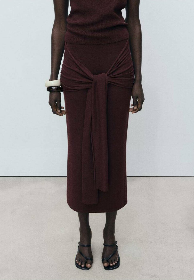 Jupe midi ajustée bordeaux avec un détail de noeud à la taille et une texture lisse. Associée à des sandales noires minimalistes.