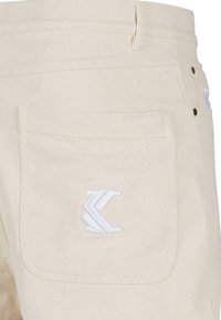 Kremfargede bomullsbukser med en stor baklomme og hvit brodert logo. Inkluderer to metallknapper og lett tekstur synlig.