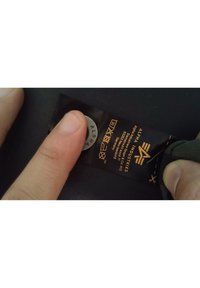 Musta kangaslabeli, jossa oranssilla tekstillä lukee "ALPHA INDUSTRIES". Hopeanvärinen ympyränmuotoinen bränditag alapuolella. Käsi painaa labelia vasten.