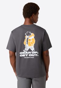 Mand iført en mørkegrå T-shirt med et grafisk motiv af en gående bjørn i en orange jakke og teksten "Gør dig klar. Kom ud. The North Face" på ryggen.