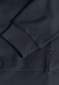 Felpa grigia scura con un tessuto testurizzato. Le caratteristiche includono una manica piegata e un logo impresso, insieme a un'etichetta piatta sul lato.