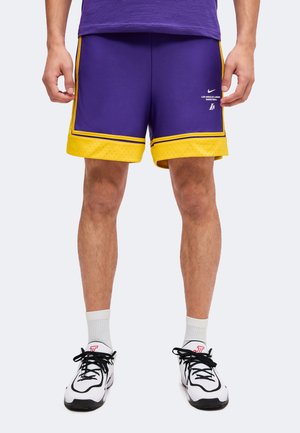 Homme portant un short de basket des Los Angeles Lakers violet et jaune, des chaussettes blanches montantes et des chaussures de sport blanches avec des lacets noirs.