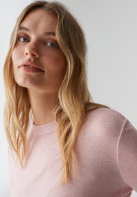 Hellrosa, geripptes Langarmshirt mit rundem Ausschnitt. Das Model hat lange Haare und einen neutralen Gesichtsausdruck vor einem minimalistischen Hintergrund.