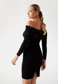 Abito nero a costine con spalle scoperte e maniche lunghe, decorato con piccoli dettagli argentati, con uno spacco alto sul retro.