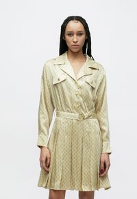 Φόρεμα με κοντά μανίκια και κουμπιά, σε ανοιχτό κίτρινο ύφασμα με επαναλαμβανόμενο γεωμετρικό σχέδιο, με ζώνη και δύο τσέπες στο στήθος με χρυσά κουμπιά.