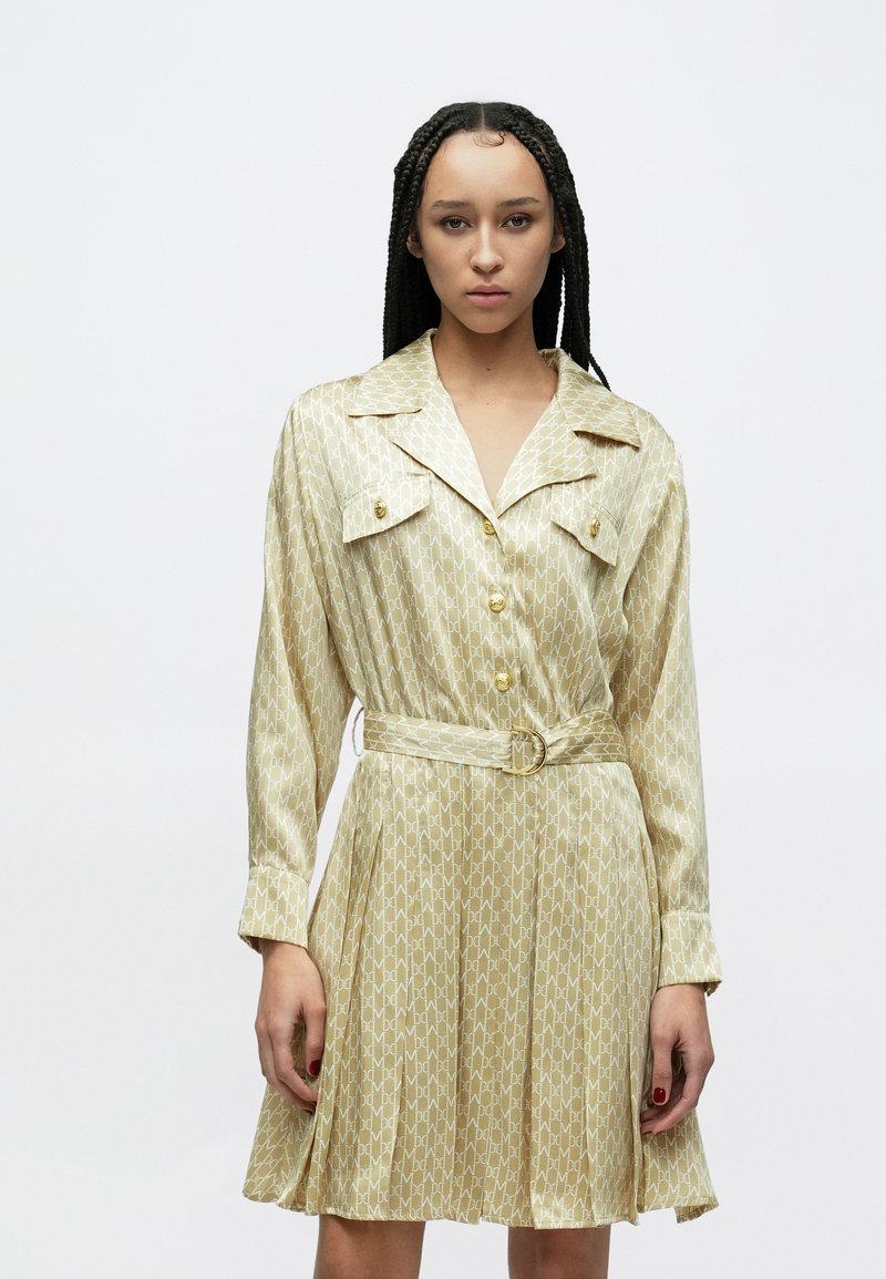 Φόρεμα με κοντά μανίκια και κουμπιά, σε ανοιχτό κίτρινο ύφασμα με επαναλαμβανόμενο γεωμετρικό σχέδιο, με ζώνη και δύο τσέπες στο στήθος με χρυσά κουμπιά.