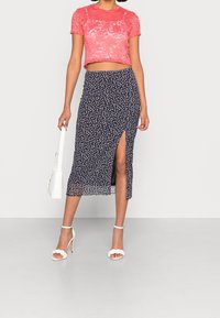 Haut court en dentelle rose, jupe midi fleurie marine avec fente, talons blancs à lanières et sac à main blanc, sur un fond uni.