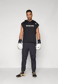 Juodas rankovių gobtuvas su užrašu „BOXING“, derinamas su juodomis kelnėmis ir baltais boksininkų pirštinėmis. Avalynė juoda su smėlio spalvos detalėmis.