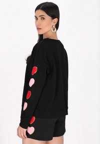 Zwarte sweatshirt met een textuurpatroon, voorzien van kleurrijke hartapplicaties in rood en roze op de mouwen. Gecombineerd met zwarte shorts.