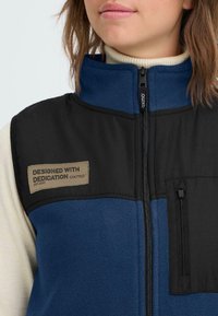 Marinblå och svart väst med fleece-textur, som har en framficka och en etikett som säger "DESIGNAD MED HENGIVENHET" på bröstet.